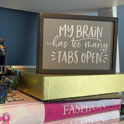 sign reads "My Brain Has Too Many Tabs Open"; the sign sits on top of a gold box on top of two books that read FASHION; in the background is a small metal peacock figurine and a blue mug with paper flowers inside