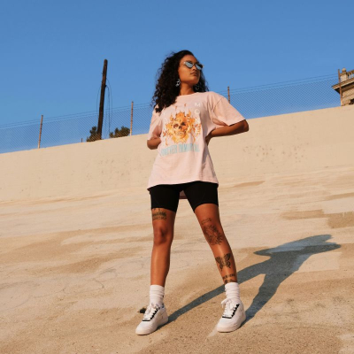 Gen Z young woman wears baggy white t-shirt, black bike shorts, white socks up to her calves, and white sneakers; she has her hands on her hips in a place with a lot of concrete (parking lot? skateboard spot?)