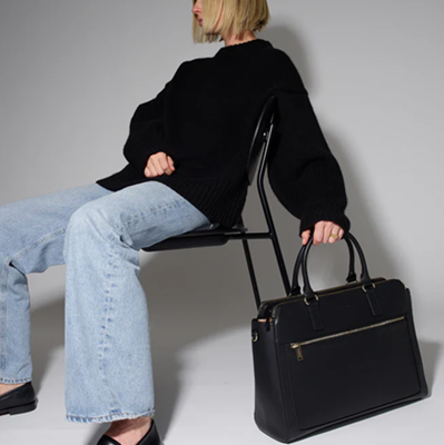 woman holds black leather work bag while seated in chair wearing black sweater and blue jeans