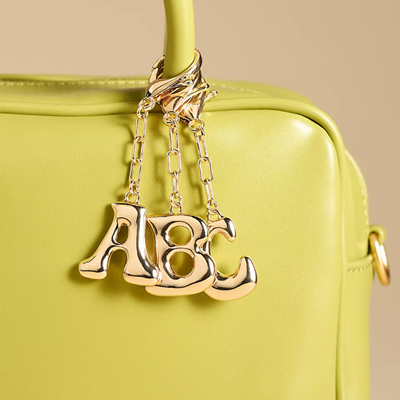 three gold bag charms hang from the strap of a bright yellow handbag; they read A, B, and C in bubble letters