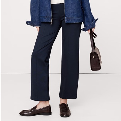 A woman wearing navy ponte pants, brown loafers, a white top, and blue denim jacket, and holding a brown croc bag