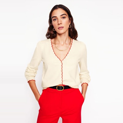A woman wearing an ivory cardigan with red scalloped trim, red pants, gold earrings, and a delicate gold necklace