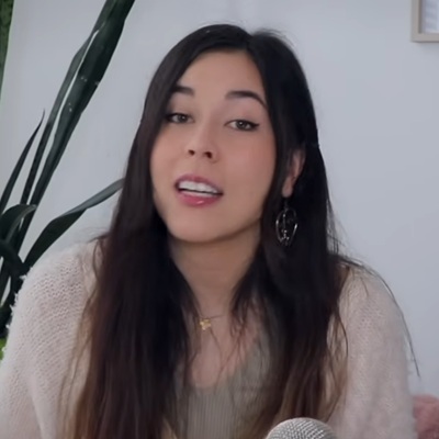 A woman talking into the camera in a YouTube video; she is wearing a gold necklace and a cream colored sweater, and there is a plant visible behind her.