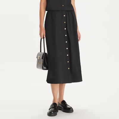 A woman wearing a black button-front skirt, black button-front top, black loafers, and carrying a black bag