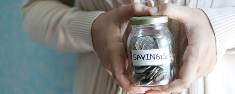 A person wearing a pinkish white cardigan holding a jar of coins marked "Savings;" she is looking for the best money tracking app
