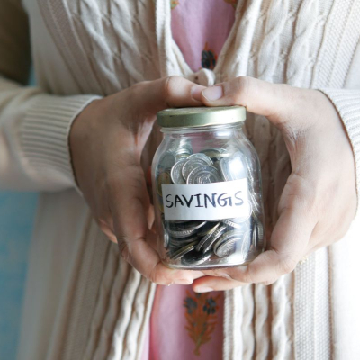 A person wearing a pinkish white cardigan holding a jar of coins marked "Savings;" she is looking for the best money tracking app