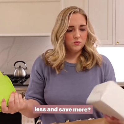 A woman in a kitchen wearing a blue shirt and holding two items; a teapot is on the stove behind her. Overlaid text says "less and save more?"