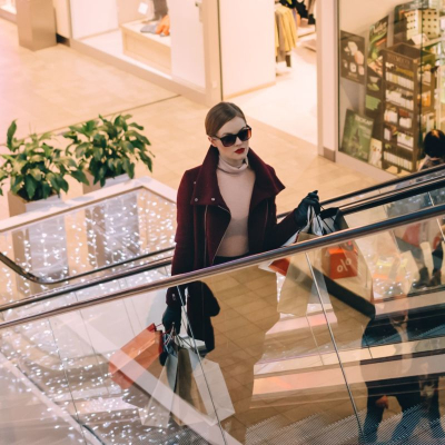 A woman on a mall escalator carrying shopping bags; she is wearing sunglasses, a brown coat, and pink top, and there are people behind her