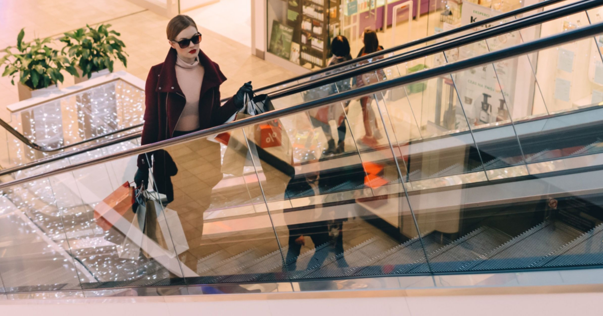 A woman on a mall escalator carrying shopping bags; she is wearing sunglasses, a brown coat, and pink top, and there are people behind her 