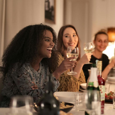 a group of women laugh together, they just might be at a book club, knitting club, cake club or other kind of club to find female friends! They are holding wine glasses and chatting over white plates; in the background you can see white walls and modern art
