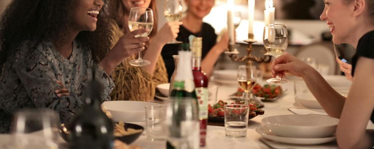 a group of women laugh together, they just might be at a book club, knitting club, cake club or other kind of club to find female friends! They are holding wine glasses and chatting over white plates; in the background you can see white walls and modern art