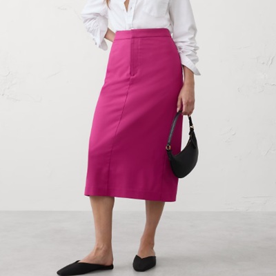 A woman wearing a pink skirt and while long sleeve blouse with black handbag and black flats