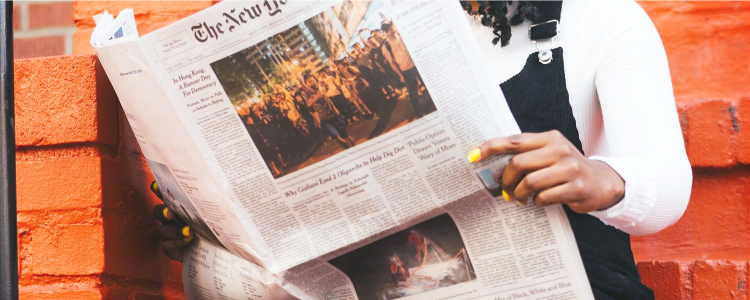 young black woman reads the NYT; she is in front of an orange brick building and has bright yellow nails; she is wearing a white shirt with a black overall