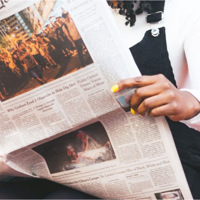 young black woman reads the NYT; she is in front of an orange brick building and has bright yellow nails; she is wearing a white shirt with a black overall