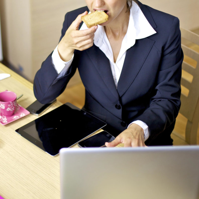 female executive eats a piece of toast while working on a computer; she is wearing a suit