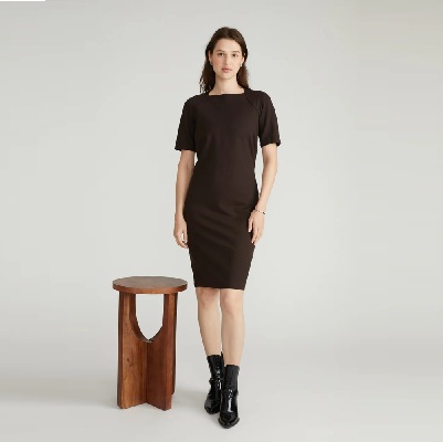 A woman wearing a brown dress and black boots, standing next to a wooden stool