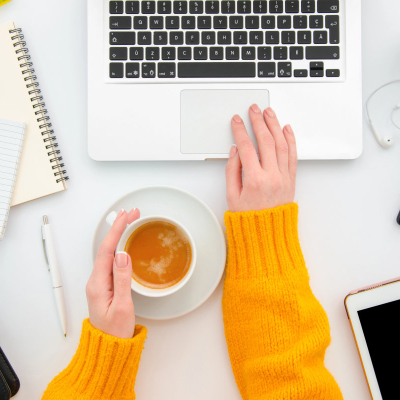 woman wearing yellow sweater types on a laptop keyboard; she is holding a cup of tea