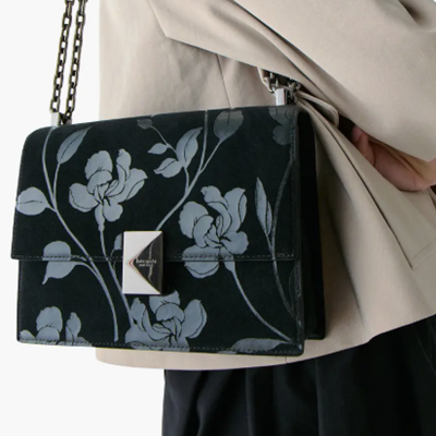 woman wears black and gray shoulder bag and a beige jacket; the shoulder bag has chain strap and a slightly pyramid-shaped closure detail