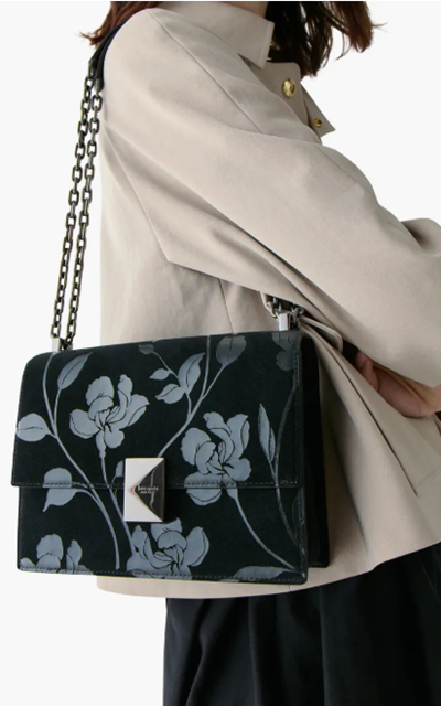 woman wears black and gray shoulder bag and a beige jacket; the shoulder bag has  chain strap and a slightly pyramid-shaped closure detail