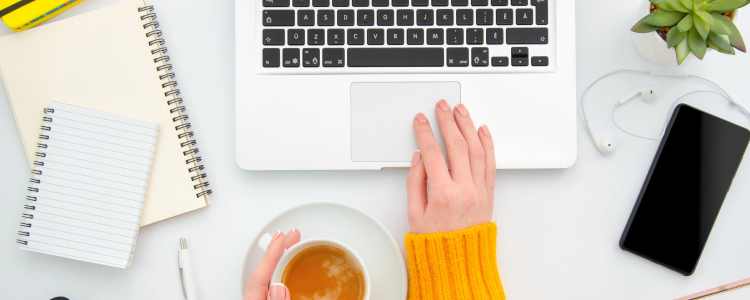 woman wearing yellow sweater types on a laptop keyboard; she is holding a cup of tea