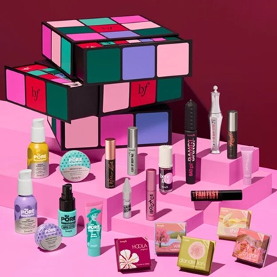 A multicolored square of cubes that is Benefit's advent calendar. It sites on a pink surface with the 24 products it includes