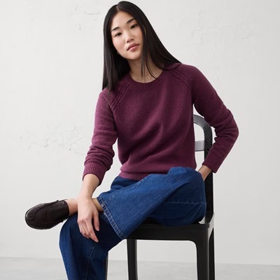 A woman with a purple sweater, blue jeans, and brown loafers. She is sitting on a black chair with her legs crossed.