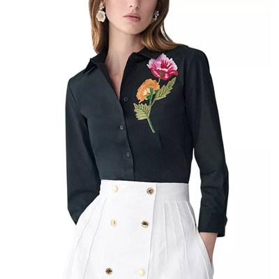 A woman wearing a black blouse with floral embroidery and a white skirt with buttons