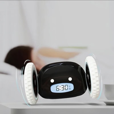 A black-and-white rolling alarm clock rolling away while a woman sleeps in a bed in the background
