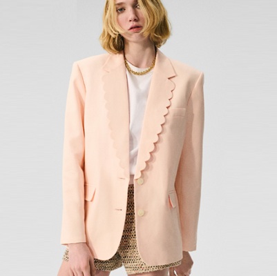 A woman wearing a peach blazer, white top, gold necklace, and brown shorts