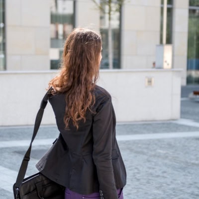 polished young businesswoman walks to work carrying a large bag; she's wearing a blazer and a purple skirt and walking along a city street