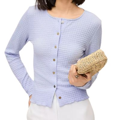 A woman wearing a light blue cardigan and white pants; she is holding a woven clutch