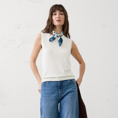 A woman wearing a white tank, white-and-blue bandana around her neck, and blue jeans. She is carrying a black jacket.