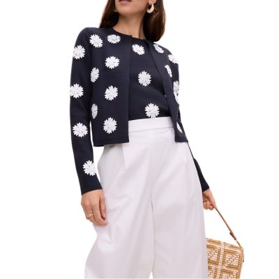 A woman wearing a navy cardigan with white floral appliques, a matching top underneath, and white wide-leg pants. She is carrying a light-brown handbag.