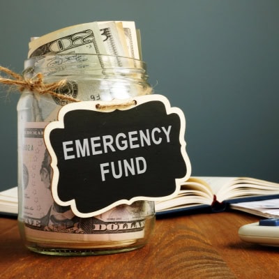 glass jar filled with money has a sign reading "emergency fund;" in the background a book is open and glasses are on a table near a calculator.