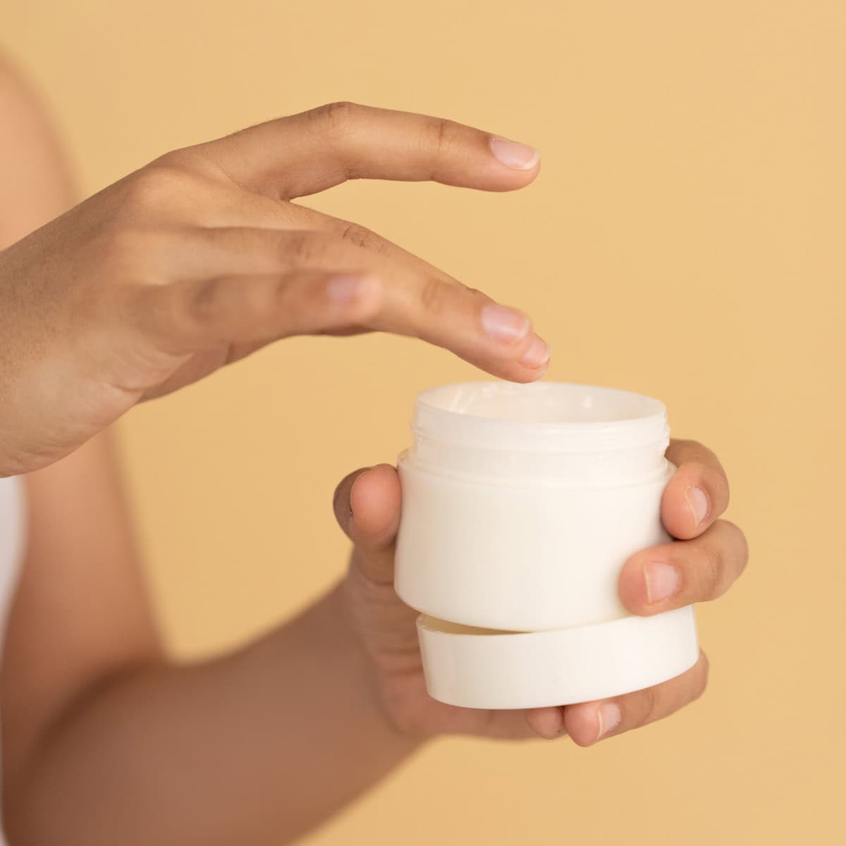 woman dips her finger into a pot of moisturizer or cream; there is a very orangey background