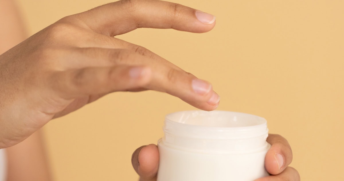 woman dips her finger into a pot of moisturizer or cream; there is a very orangey background