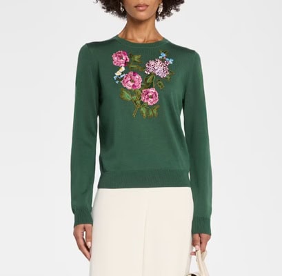 A woman wearing a dark green sweater embroidered with flowers, and ivory pants