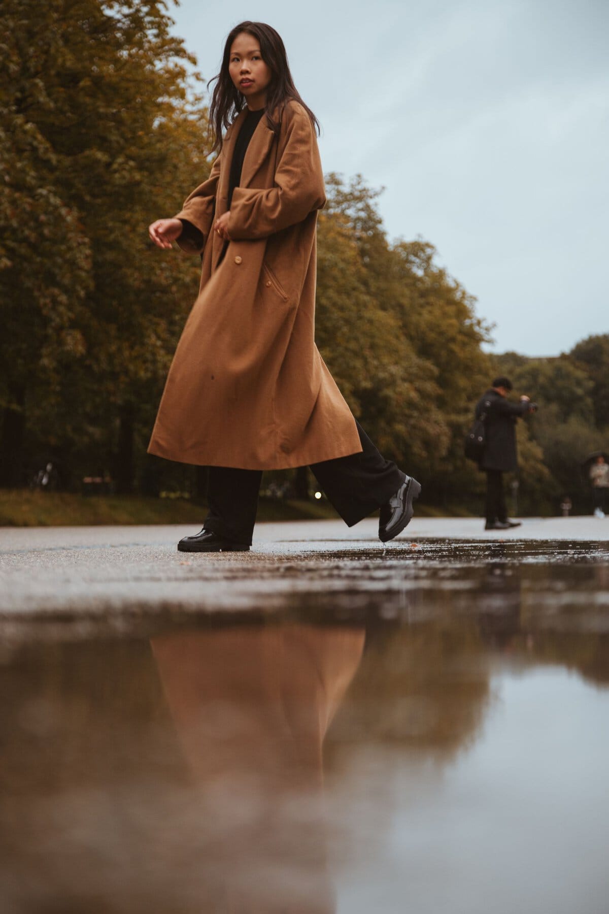A woman in a brown coat walks through a park in Munich, reflecting in a puddle, on a cloudy day.