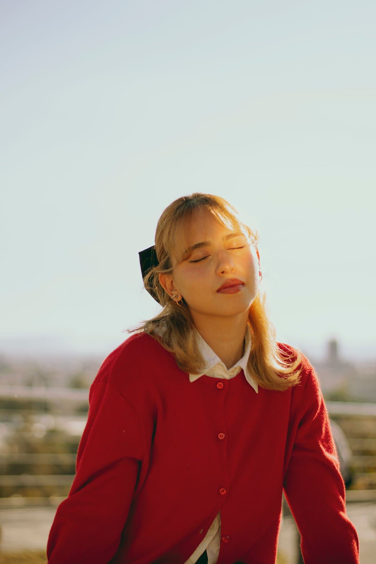 A young woman in a red sweater basks in warm sunlight outdoors, eyes closed.