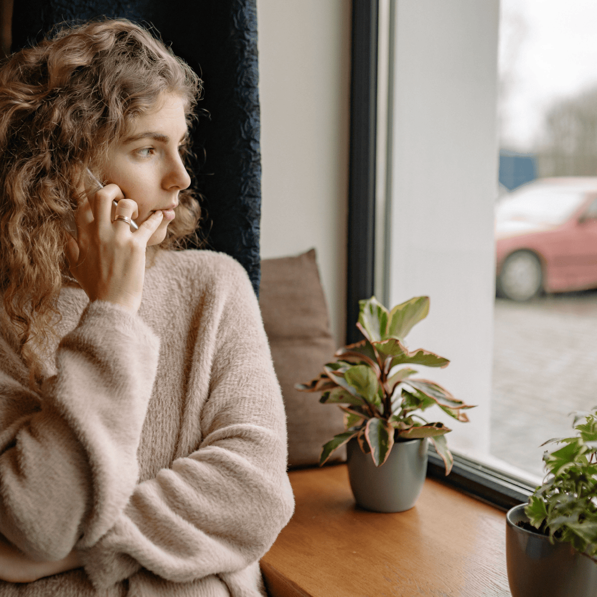 woman looks out window; she is wearing a beige sweater and has a contemplative expression on her face