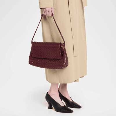 woman holds burgundy bag with woven leather detail; she is wearing weird heels and a beige dress or trench