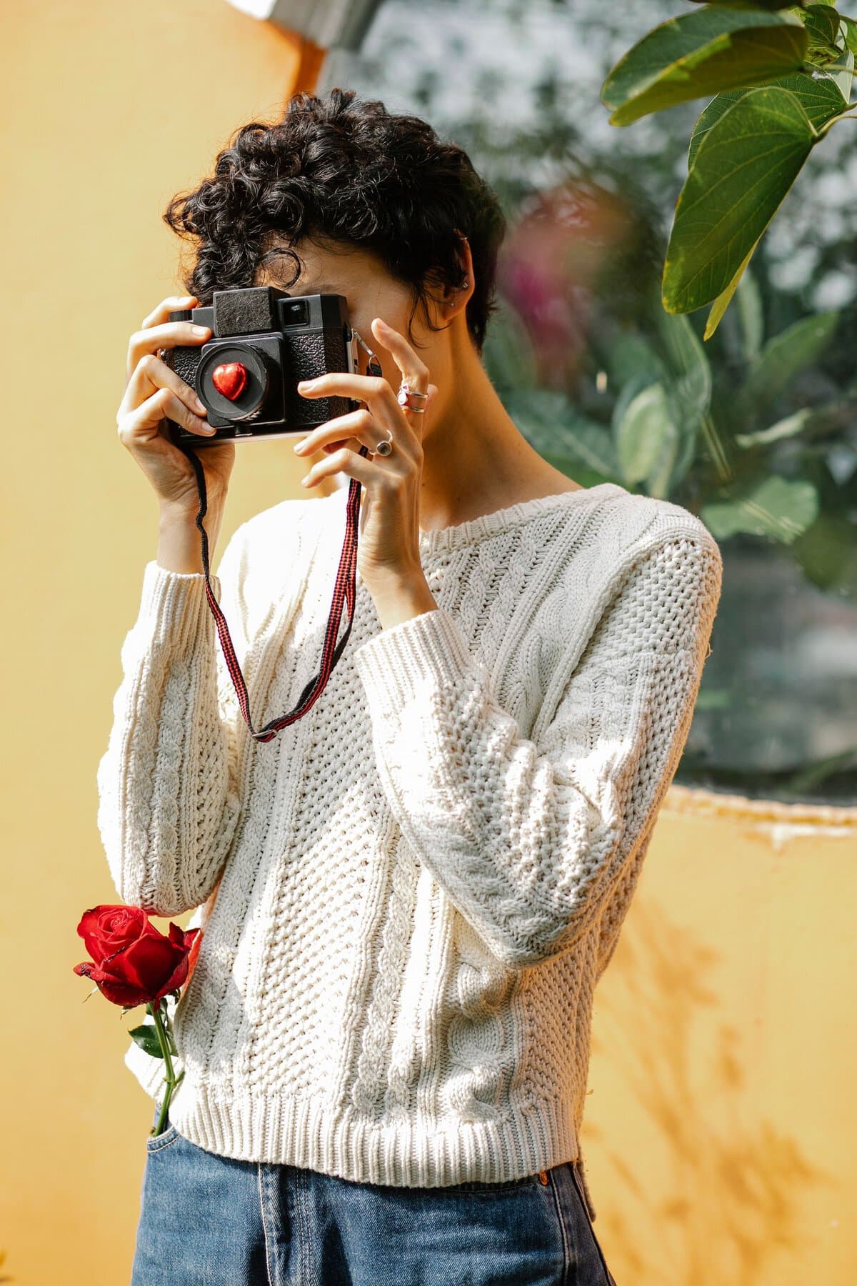 Faceless focused female photographer in casual wear and red rose in pocket taking picture on photo camera while standing near wall