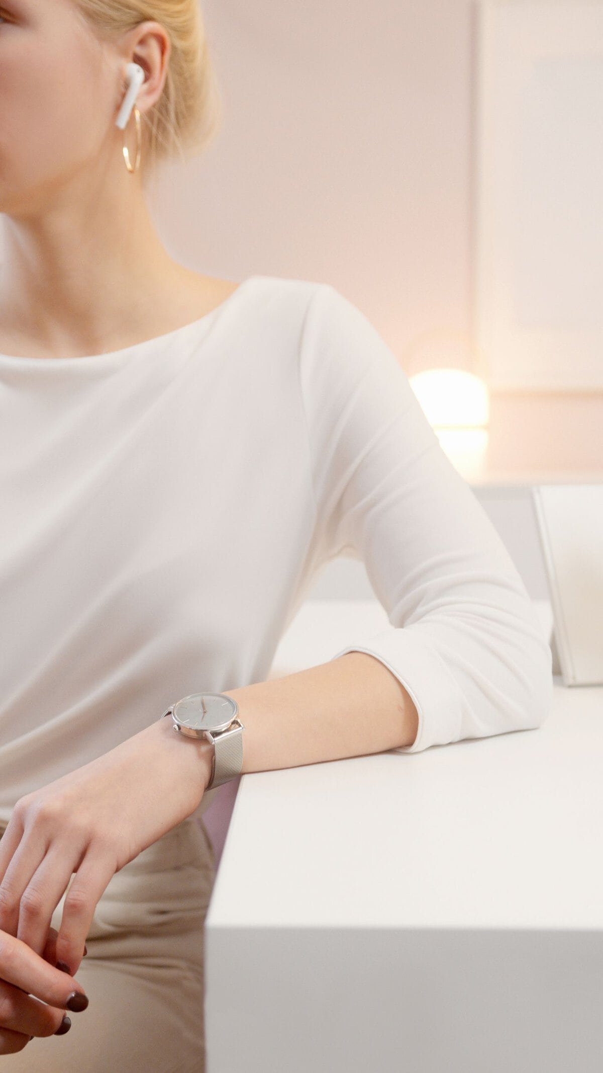Sophisticated woman wearing earbuds and wristwatch, sitting elegantly indoors with modern lighting.