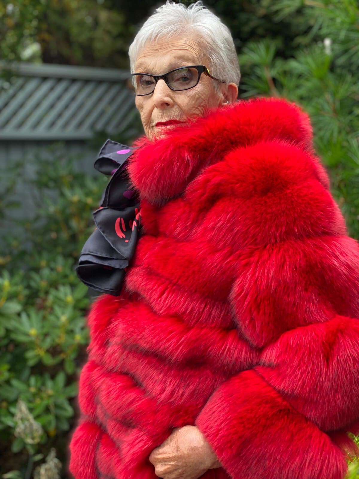 Stylish senior woman wearing a vibrant red fur coat and sunglasses in a garden setting.