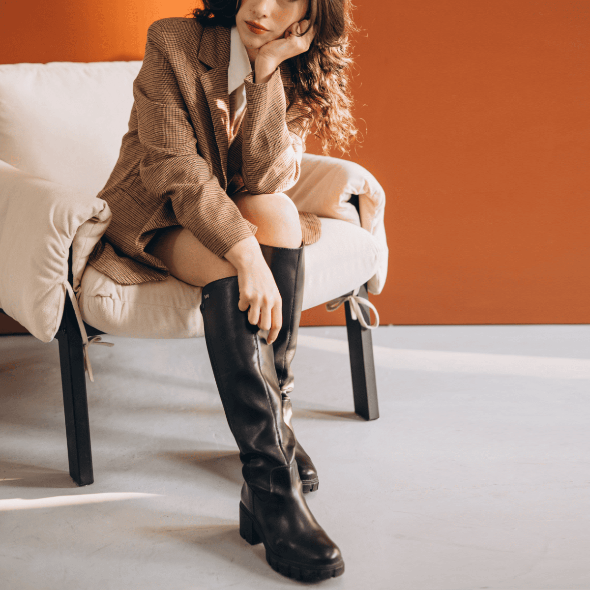 professional woman lounges in a chair; she is wearing black knee-high boots with bare legs, a shorter skirt, and a blazer