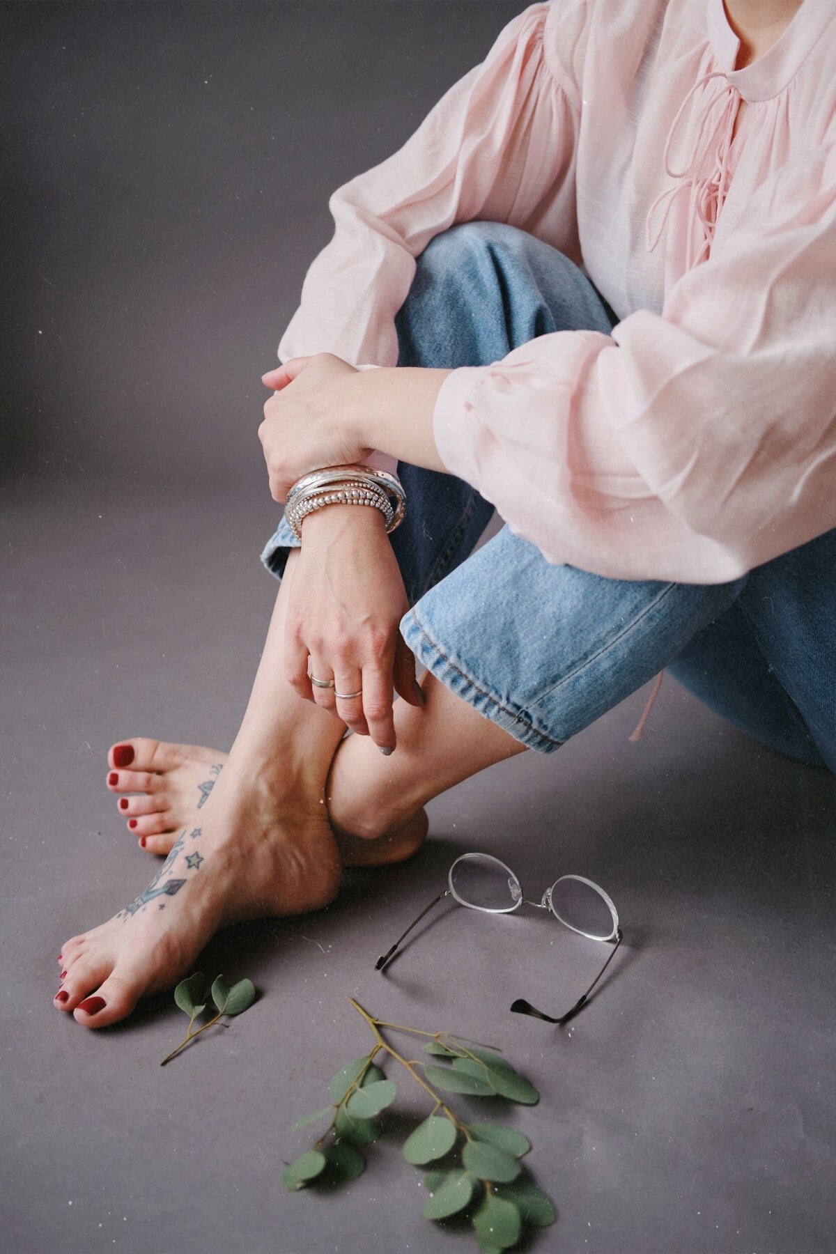 Woman sitting barefoot indoors with eyeglasses and leaves around her. Fashion-forward and relaxed style.
